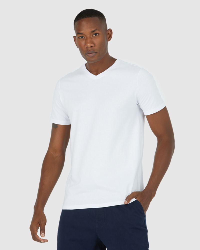 Homem negro e cabelo preto com mão no bolso utilizando camiseta branca básica masculina gola V em algodão e calça azul.