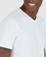 Homem negro e cabelo preto com mão no bolso utilizando camiseta branca básica masculina gola V em algodão e calça azul.