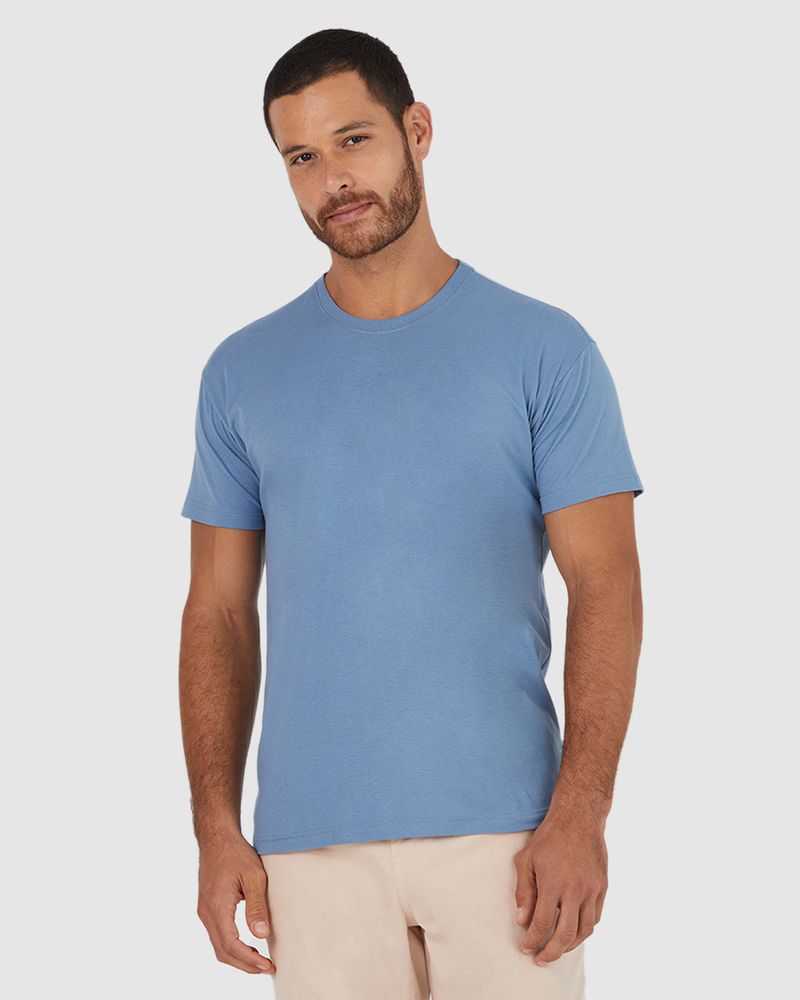 Homem branco de cabelo preto e mão no bolso utilizando camiseta básica azul claro gola texturizada em algodão.