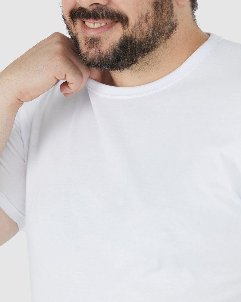 Homem branco de cabelo preto sorrindo e utilizando camiseta básica plus size masculina cor branca com decote redondo em algodão e bermuda cinza.