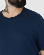 Homem branco de cabelo preto sorrindo e utilizando camiseta básica plus size, cor azul escuro masculina com decote redondo em algodão e calça jeans.